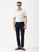 A man wearing a short-sleeve polo shirt in off-white with navy blue and gold trim on the collar and sleeve cuffs, paired with dark navy trousers, against a plain light gray background.
