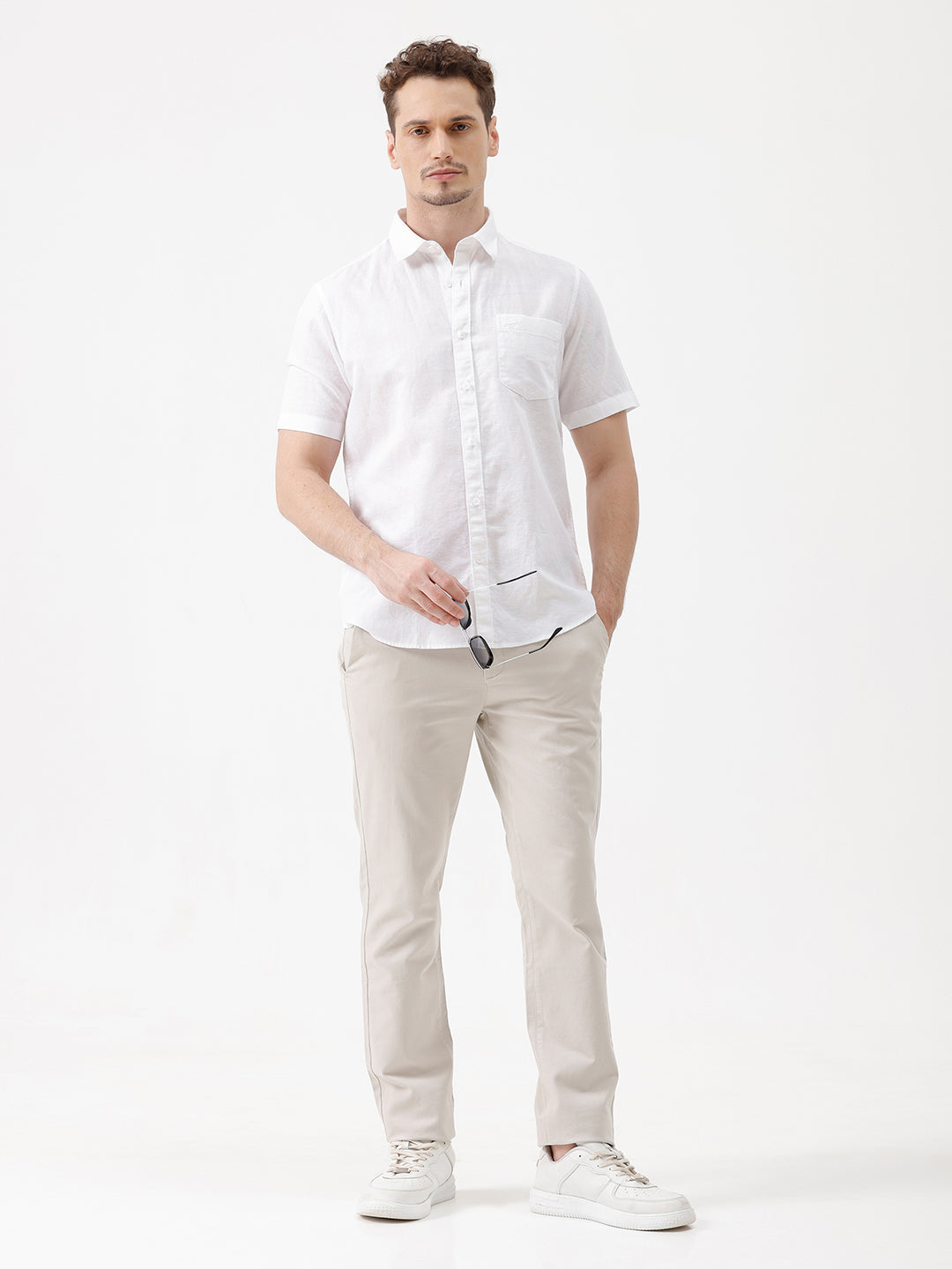 A man wearing a white short-sleeve button-up shirt with a chest pocket and beige trousers, standing against a plain white studio background.