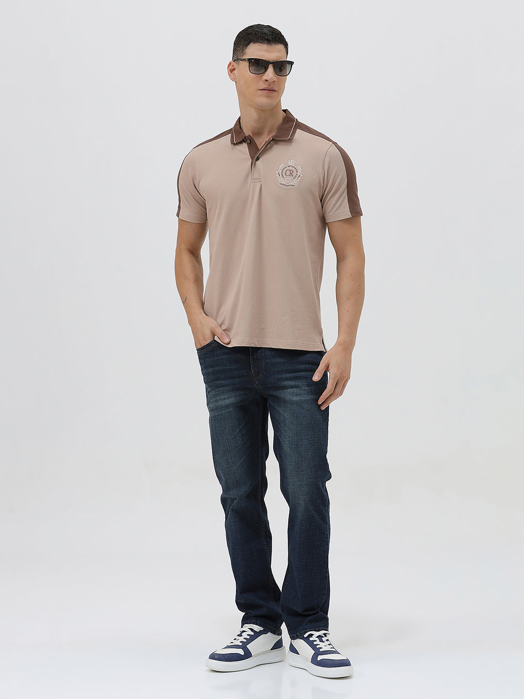 A man modeling a short-sleeve polo shirt in beige with brown shoulder panels and collar trim, paired with dark blue jeans, against a light gray studio background.