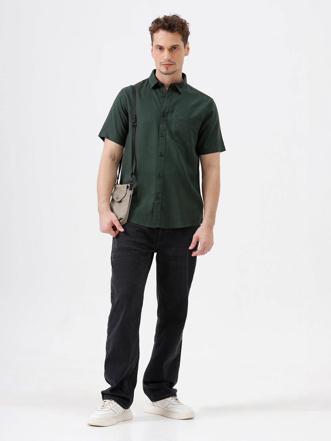 A man wearing a dark forest green short-sleeve button-up shirt with a chest pocket, paired with black jeans, standing against a plain white studio background.
