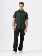 A man wearing a dark forest green short-sleeve button-up shirt with a chest pocket, paired with black jeans, standing against a plain white studio background.