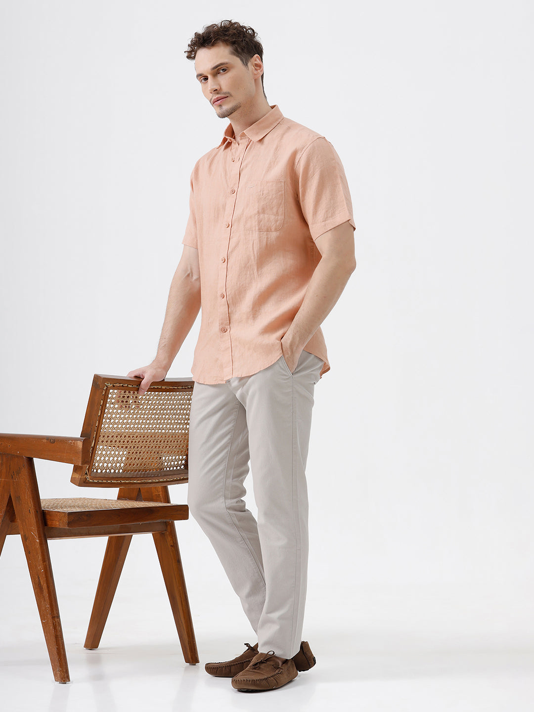 A man wearing a short-sleeve linen button-up shirt in a light peach-pink color, paired with light gray trousers, standing against a plain light gray background.