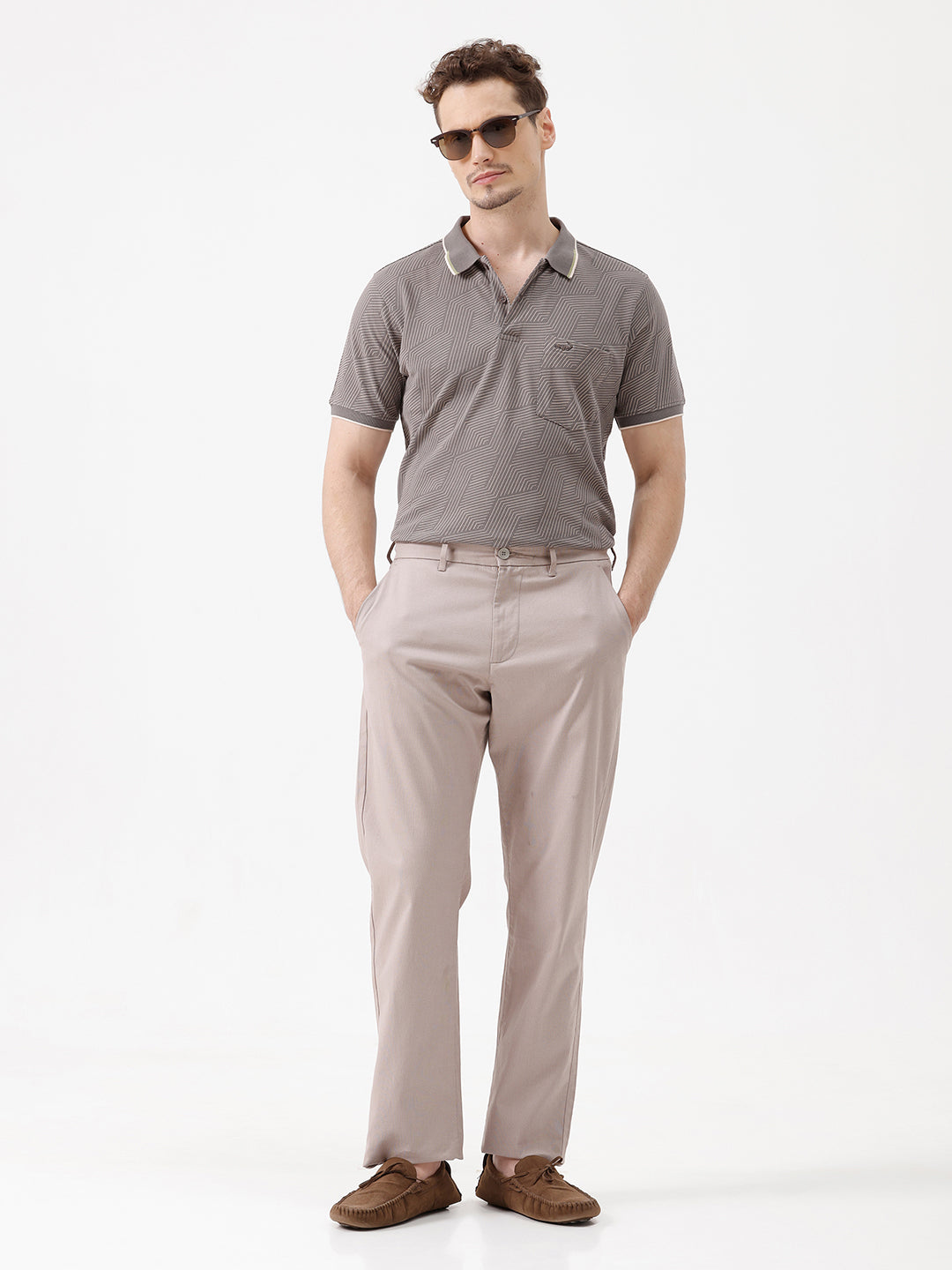 A man wearing a short-sleeve polo shirt with a gray and white geometric pattern, paired with beige trousers, standing against a plain white studio background.