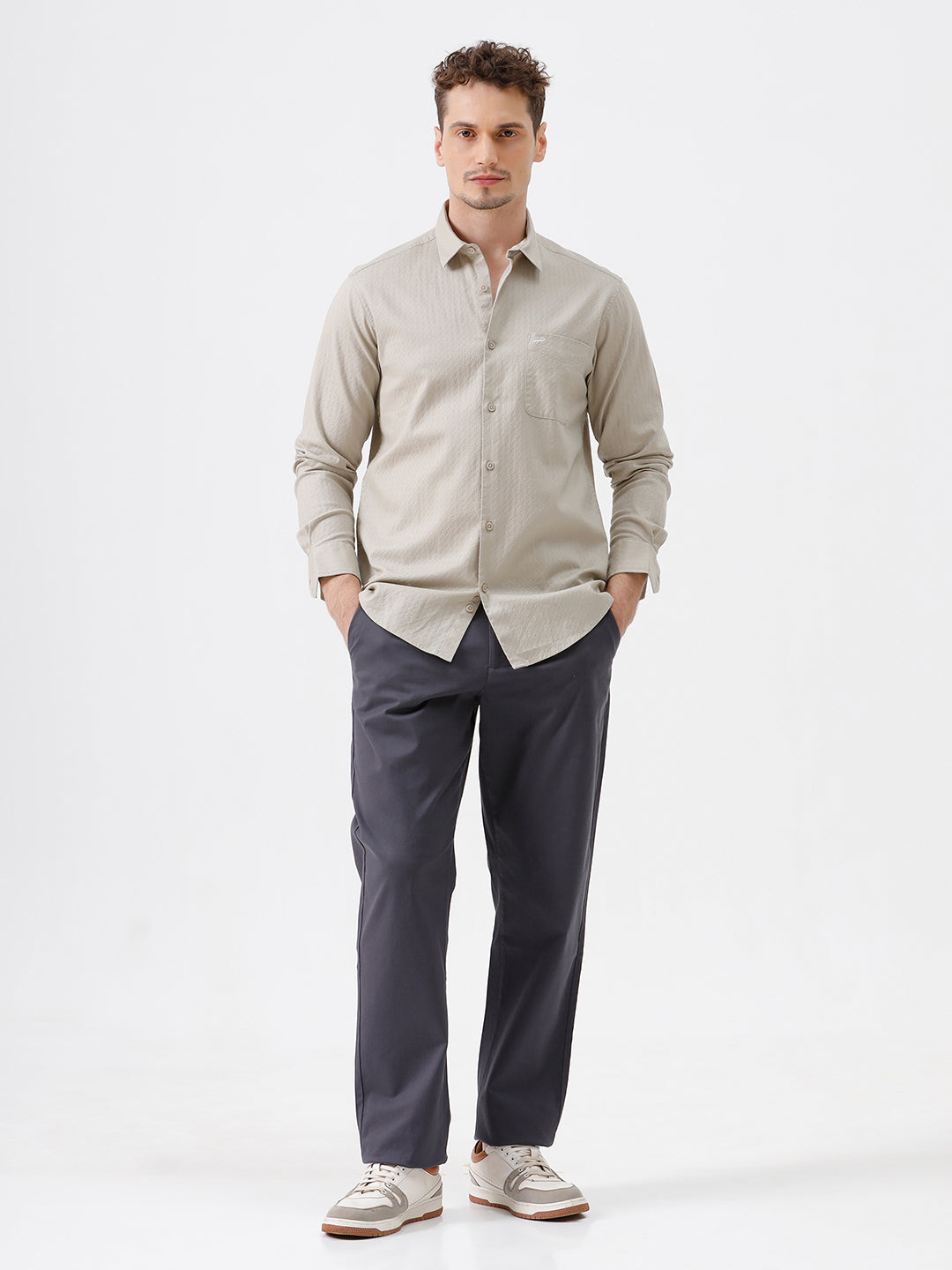 A man wearing a light beige long-sleeve button-down shirt with a chest pocket, paired with dark gray trousers, standing against a plain white studio background.