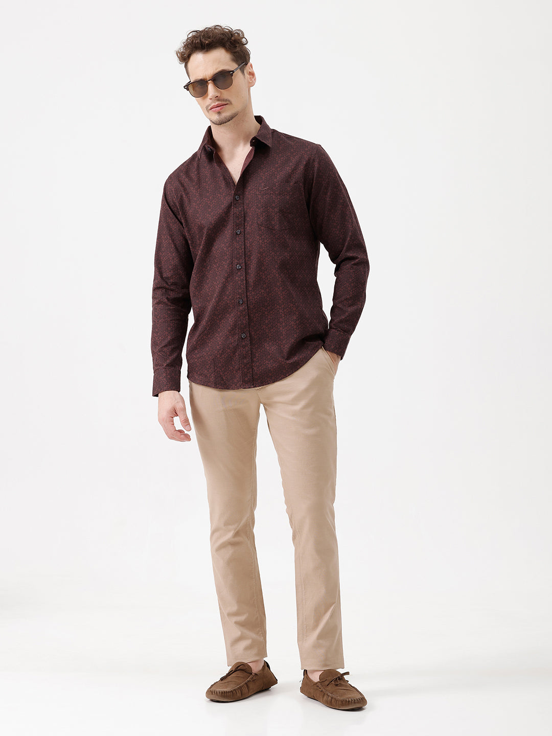 A man wearing a dark burgundy long-sleeve button-up shirt with a subtle geometric pattern, paired with beige trousers, standing against a plain white studio background.