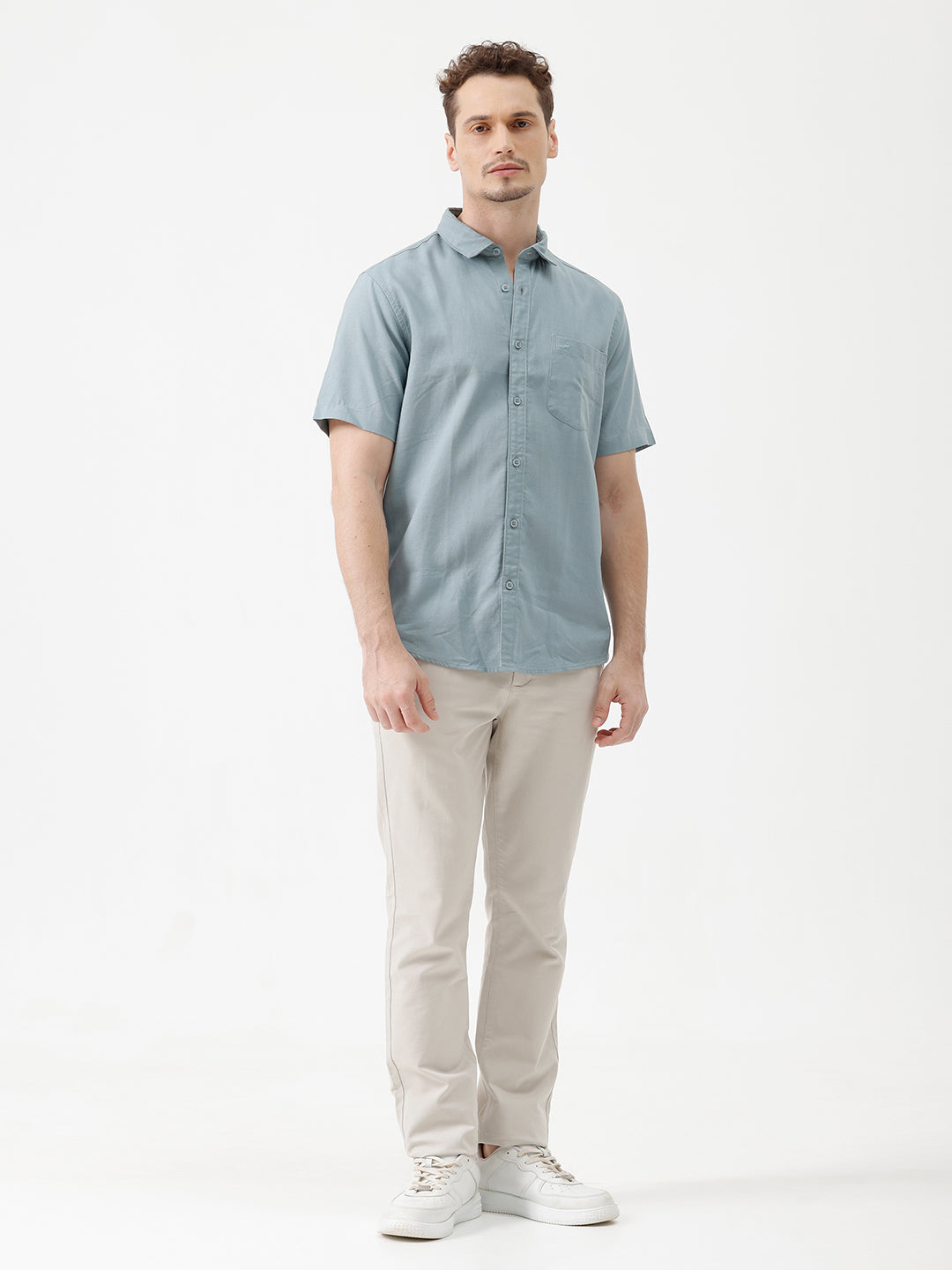 A man wearing a light blue short-sleeve button-up shirt and beige trousers, standing against a plain white studio background.