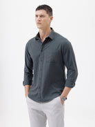 A man wearing a dark charcoal gray long-sleeve button-down shirt with a chest pocket, paired with light gray trousers, standing against a plain white studio background.
