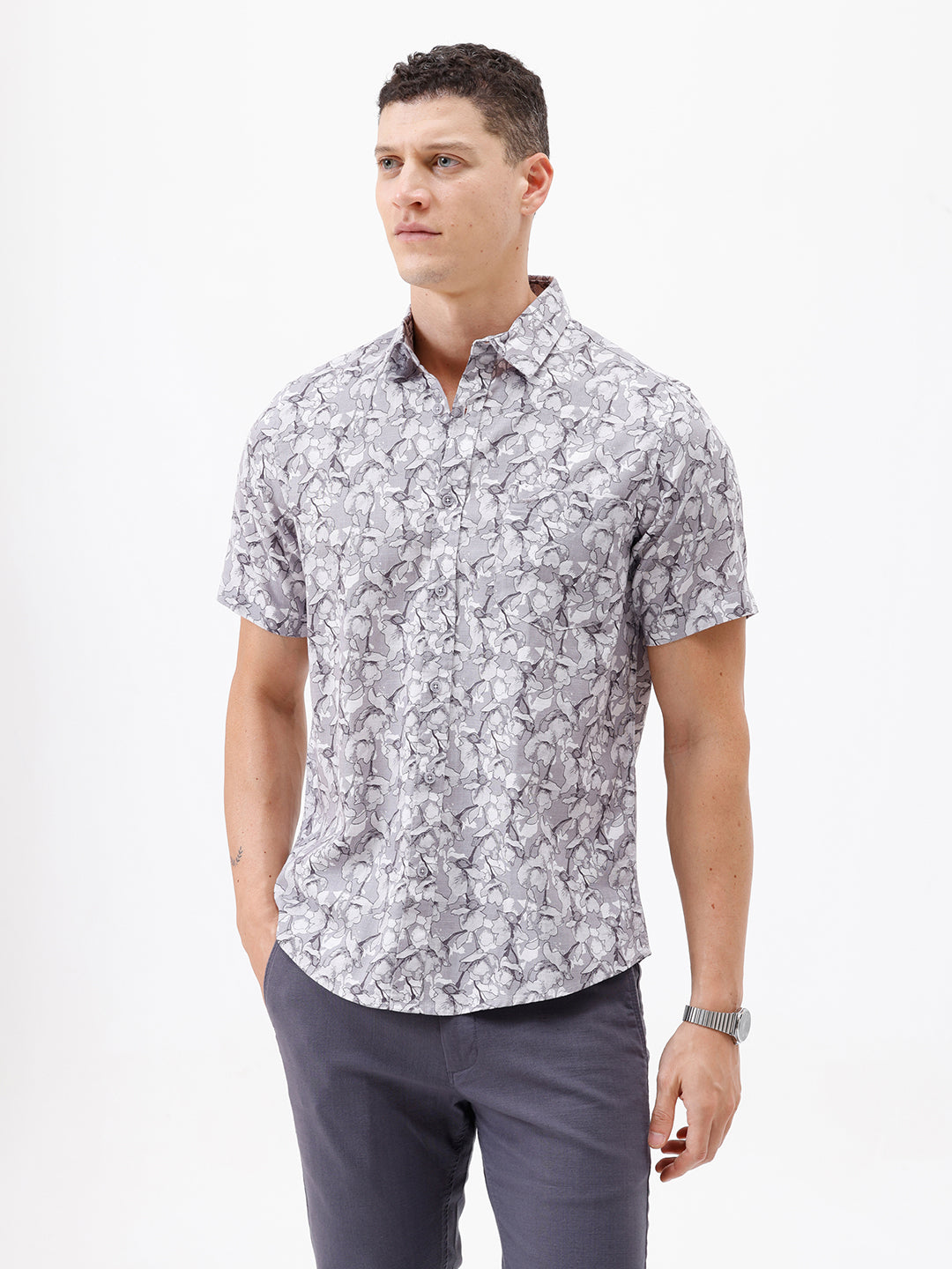A man wearing a short-sleeve button-up shirt with a gray and white abstract pattern, paired with dark gray trousers, standing against a plain white studio background.
