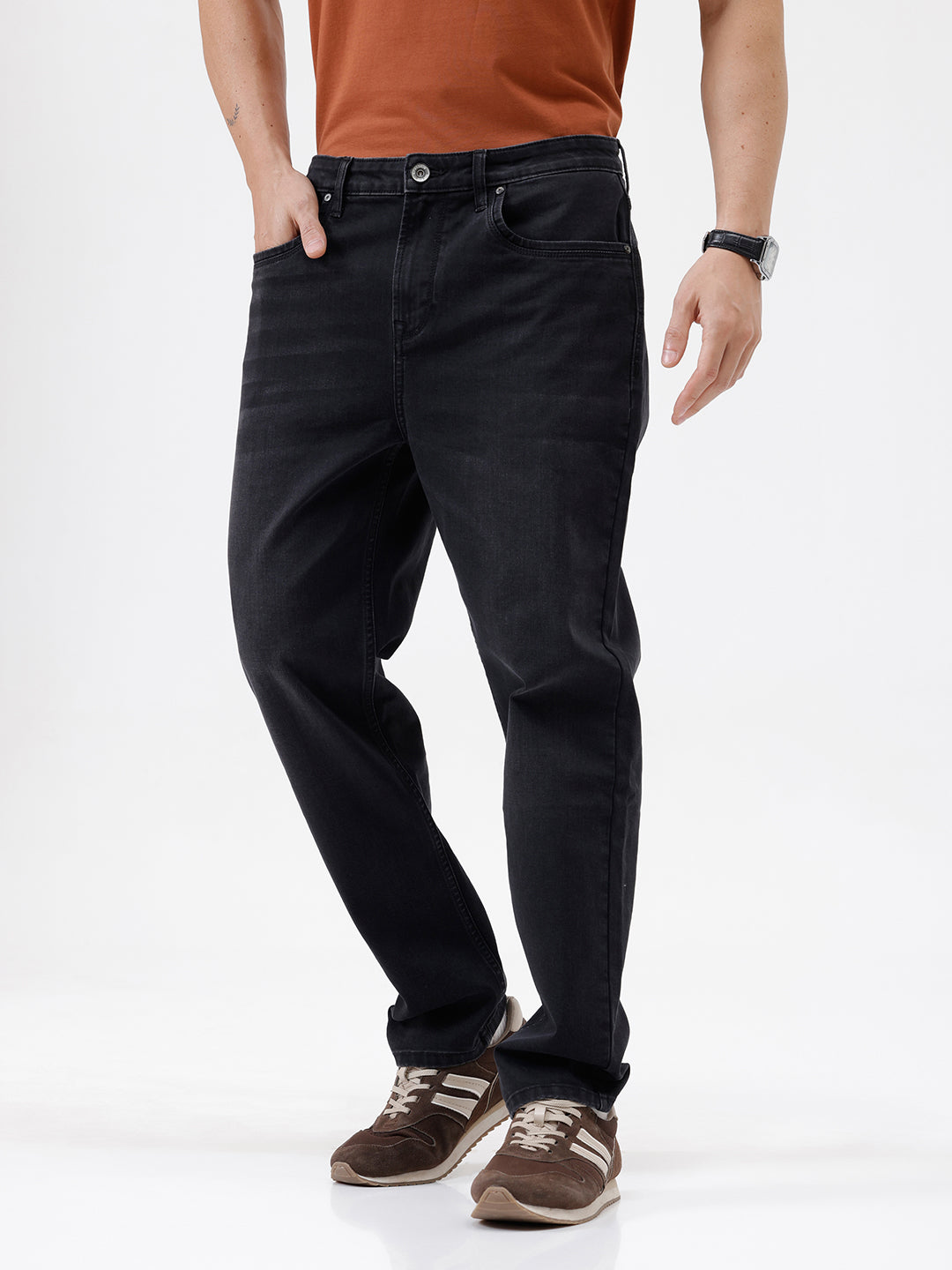 A person wearing black slim-fit jeans, paired with a rust-orange t-shirt and brown sneakers, shown from the waist down against a plain white studio background.