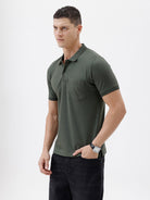A man wearing a dark olive green short-sleeve polo shirt with a chest pocket, paired with dark jeans, standing against a plain light gray background.
