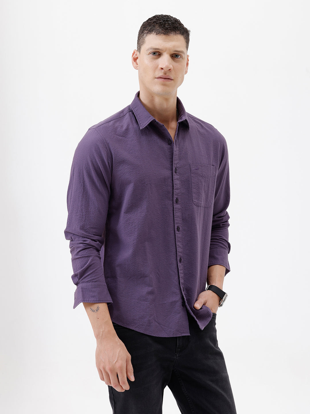 A man wearing a long-sleeve button-down shirt in a muted purple color, paired with black pants, standing against a plain white studio background.
