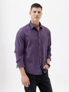 A man wearing a long-sleeve button-down shirt in a muted purple color, paired with black pants, standing against a plain white studio background.
