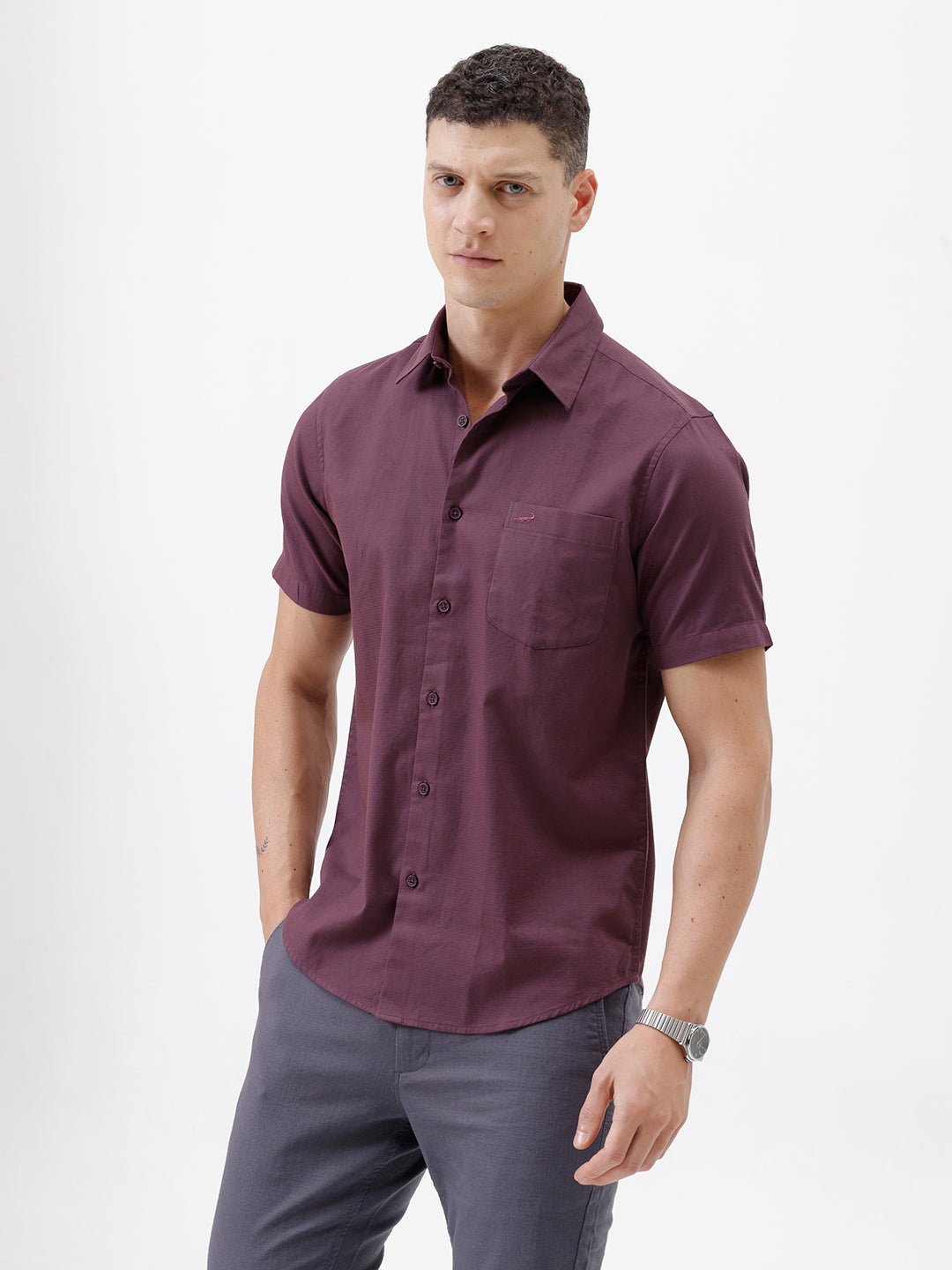 A man modeling a short-sleeve button-up shirt in a deep plum purple color, paired with gray trousers, against a plain white studio background.