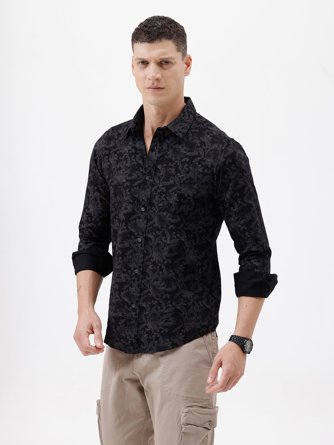 A man wearing a black long-sleeve button-up shirt with a subtle tonal floral pattern, paired with beige cargo pants and dark sunglasses, standing against a plain white background.