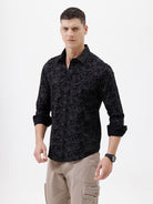 A man wearing a black long-sleeve button-up shirt with a subtle tonal floral pattern, paired with beige cargo pants and dark sunglasses, standing against a plain white background.