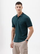 A man wearing a dark teal short-sleeve polo shirt with a chest pocket, paired with beige cargo pants, standing against a plain white studio background.