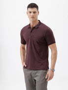 A man wearing a short-sleeve polo shirt in dark burgundy with a chest pocket, paired with gray trousers, standing against a plain light gray background.
