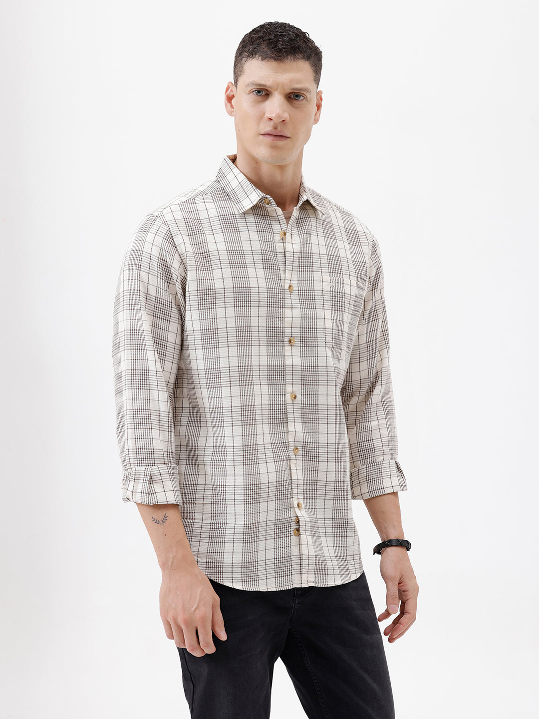 A man wearing a long-sleeve button-up plaid shirt in beige, white, and gray, paired with dark pants, standing against a plain white studio background.