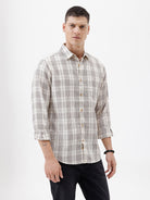 A man wearing a long-sleeve button-up plaid shirt in beige, white, and gray, paired with dark pants, standing against a plain white studio background.
