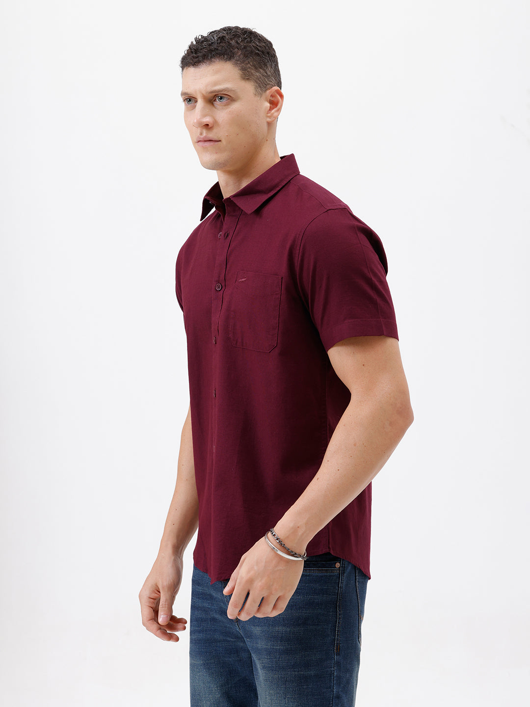 A man wearing a short-sleeve button-up shirt in maroon with a chest pocket, paired with blue jeans, standing against a plain white studio background.