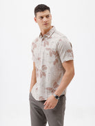 A man wearing a short-sleeve button-up shirt with a beige and brown floral pattern, paired with gray trousers, standing against a plain white background.