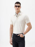 A man wearing a short-sleeve polo shirt in cream with subtle horizontal textured stripes, paired with dark jeans, standing against a plain white studio background.
