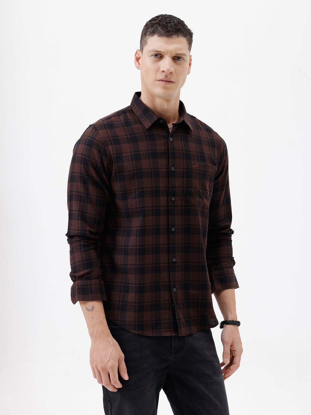 A man wearing a dark brown and black plaid long-sleeve button-up shirt, paired with dark pants, standing against a plain light gray background.