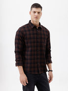 A man wearing a dark brown and black plaid long-sleeve button-up shirt, paired with dark pants, standing against a plain light gray background.