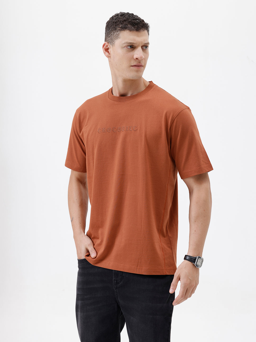 A man wearing a rust-orange crewneck t-shirt with a subtle tonal 'ELECTRIC' logo across the chest, paired with black pants, photographed against a clean white studio background.