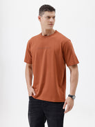A man wearing a rust-orange crewneck t-shirt with a subtle tonal 'ELECTRIC' logo across the chest, paired with black pants, photographed against a clean white studio background.