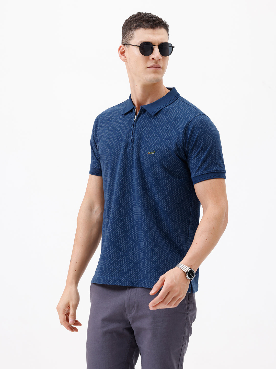 A man wearing a navy blue short-sleeve polo shirt with a subtle geometric pattern and a half-zip collar, paired with gray trousers, against a plain white background.