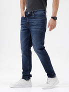 A person wearing dark blue slim-fit jeans with a faded wash, paired with a black t-shirt and white sneakers, shown from the waist down against a plain white studio background.