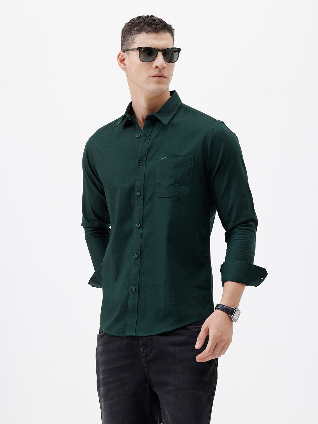 A man wearing a dark forest green long-sleeve button-down shirt with a chest pocket, paired with dark jeans, standing against a plain white studio background.