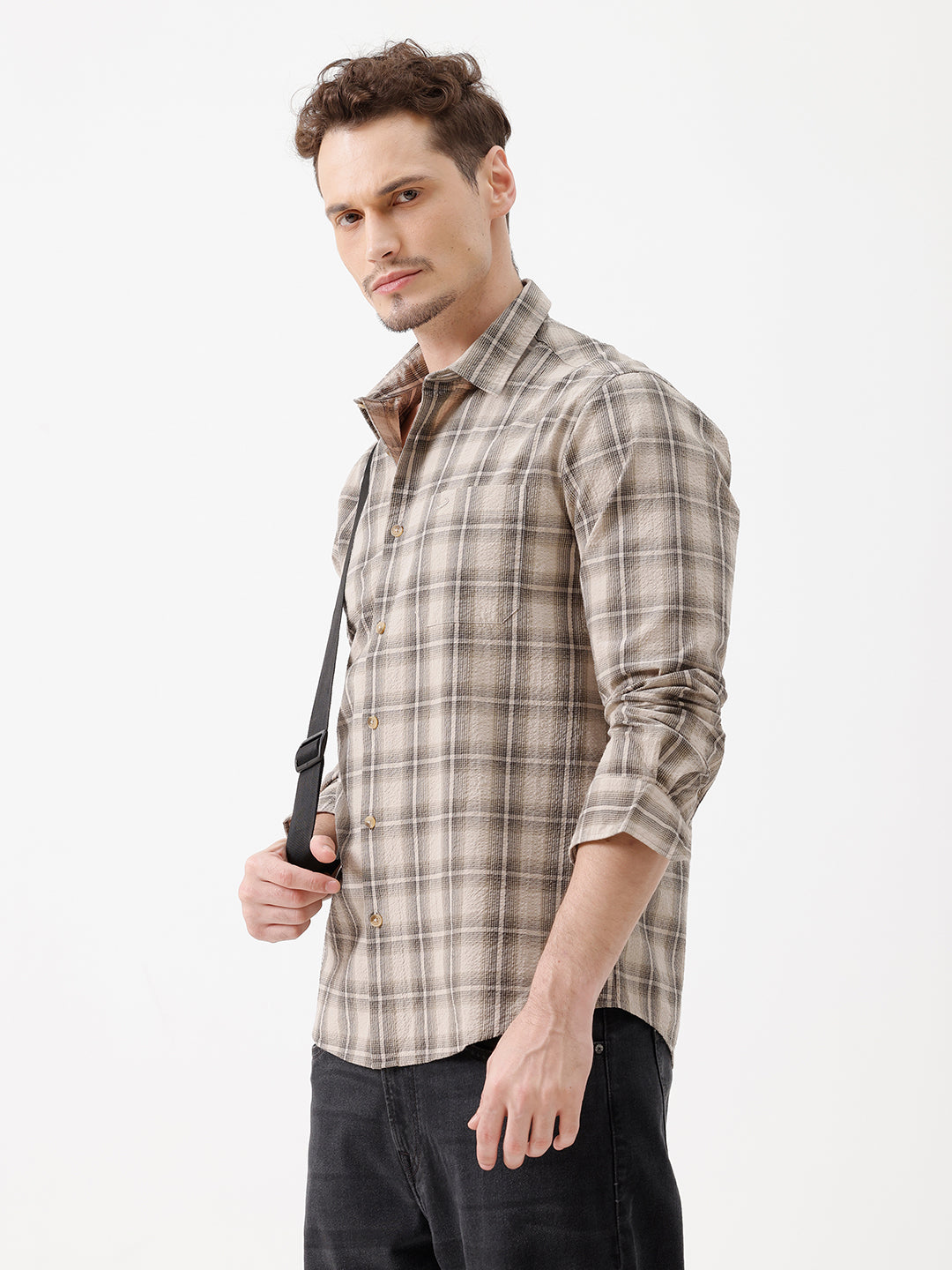 A man wearing a long-sleeve button-up plaid shirt in beige and brown tones, paired with dark jeans, standing against a plain white studio background.