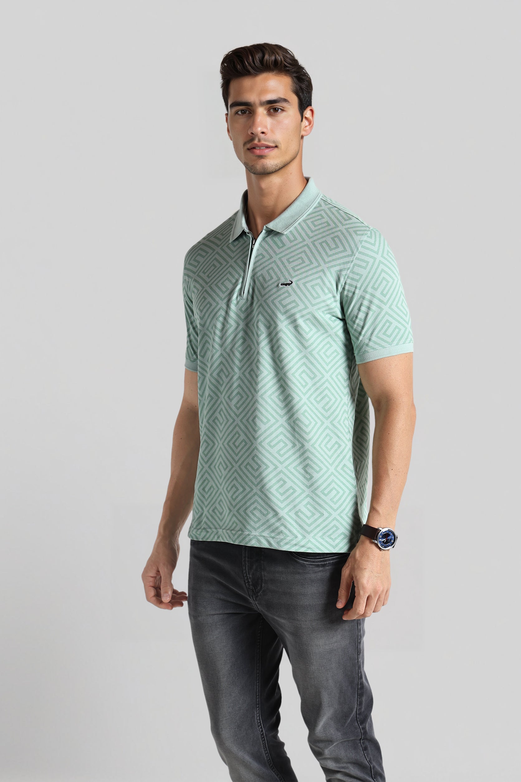 Man wearing a light green patterned polo shirt and dark jeans on a gray background