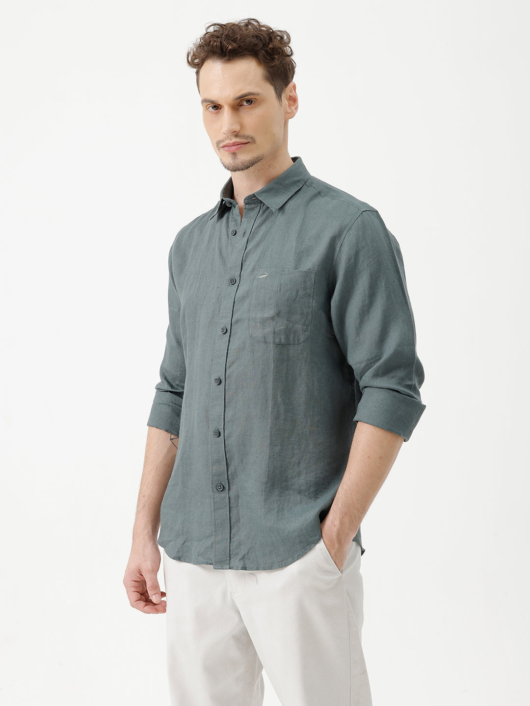 A man wearing a slate blue-green long-sleeve linen button-up shirt with white trousers, standing against a plain white studio background.