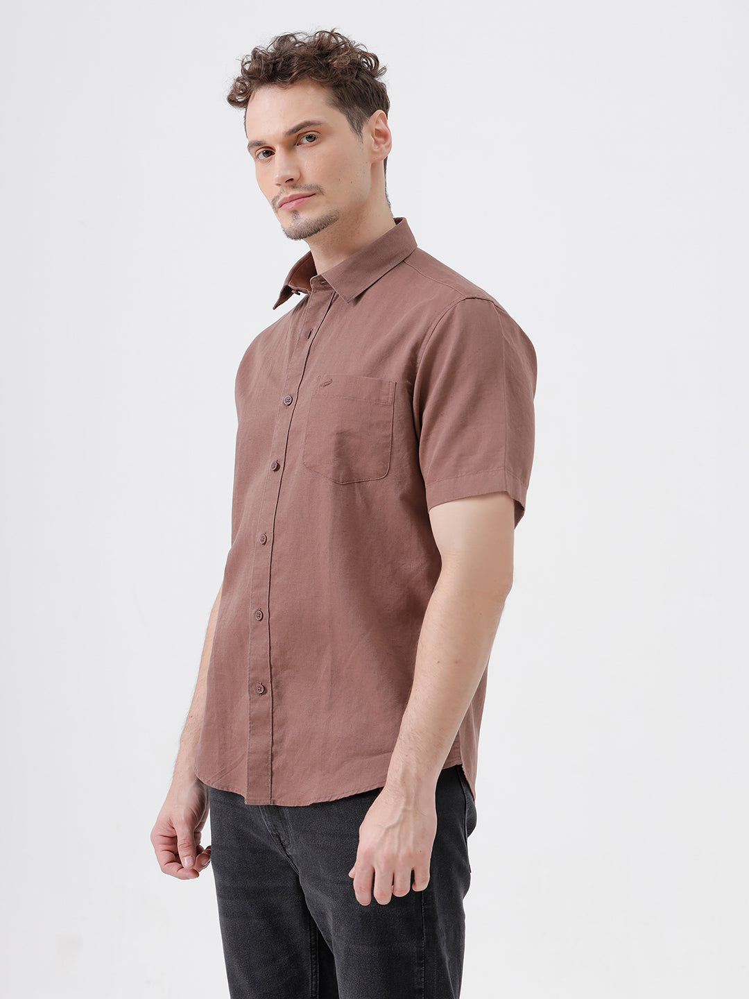 A man wearing a short-sleeve button-up shirt in a dusty rose-brown color, paired with black jeans, standing against a plain light gray background.