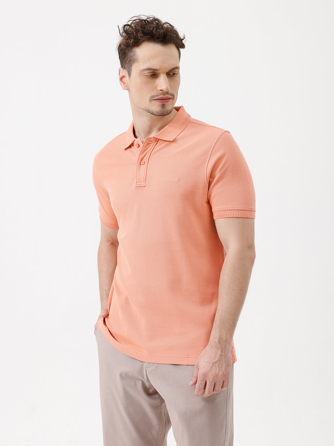 A man wearing a peach-colored short-sleeve polo shirt and beige trousers, standing against a plain white studio background.