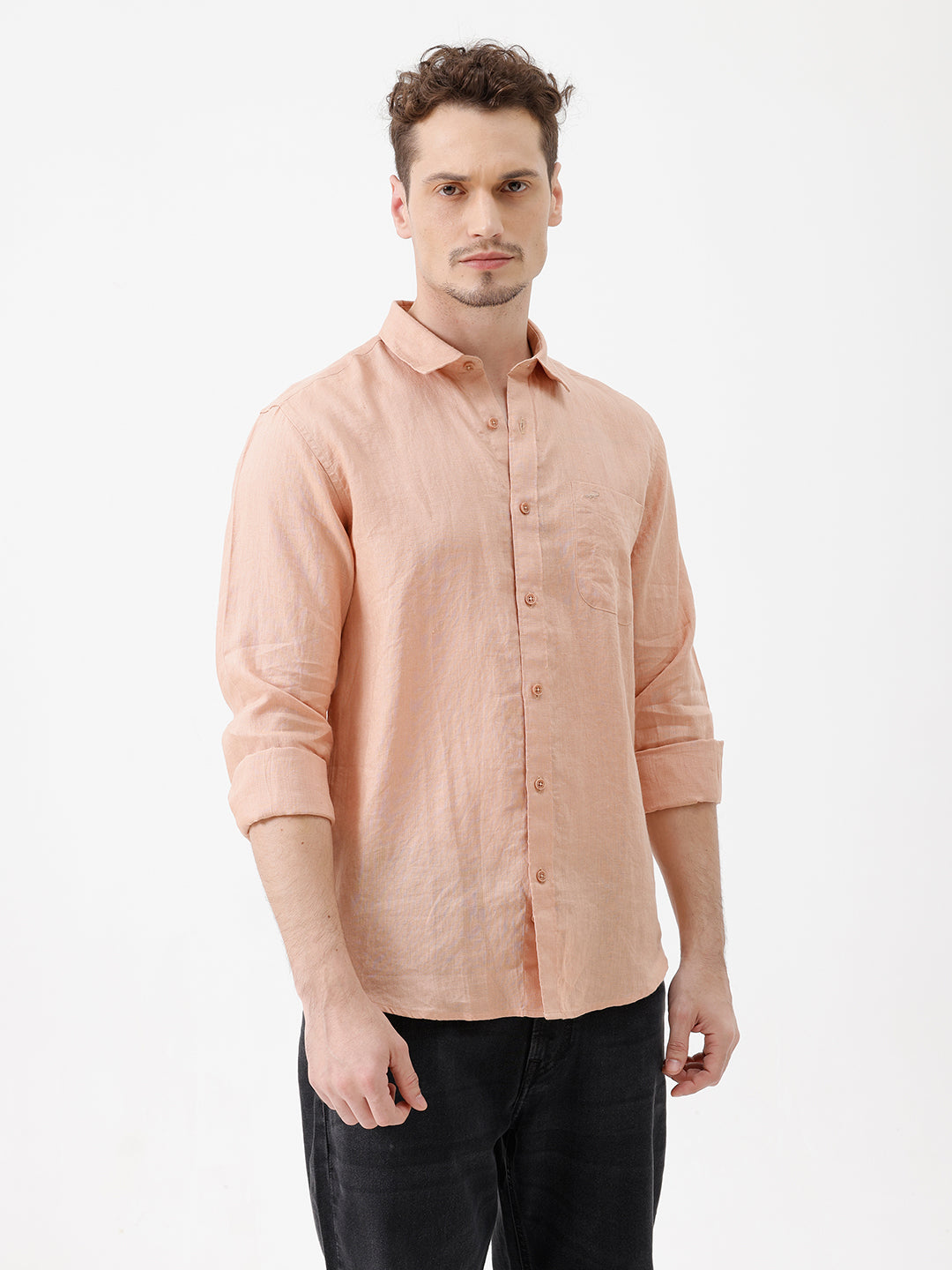 A man wearing a light peach-colored linen button-down shirt with dark jeans, standing against a plain white studio background.
