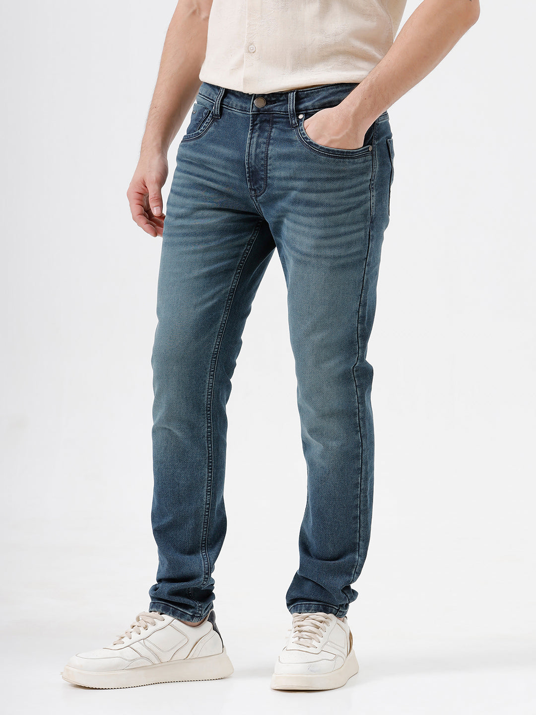 A person wearing slim-fit medium wash blue jeans, a cream-colored short-sleeve button-up shirt, and white sneakers, standing against a plain white studio background.