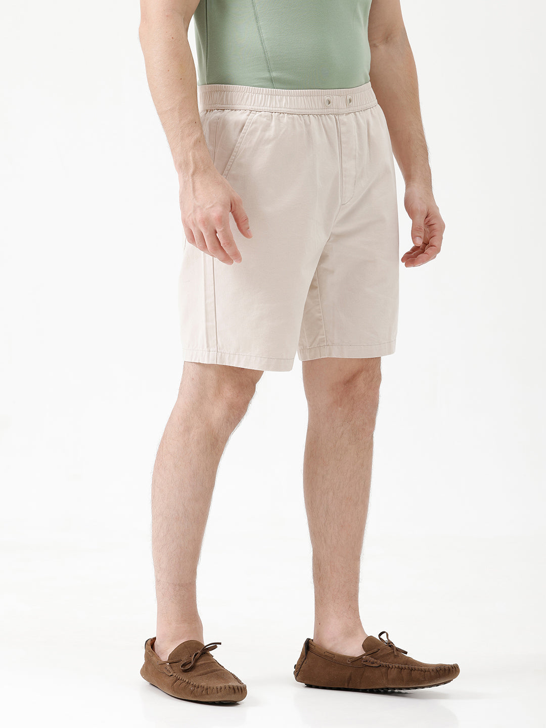 A man wearing light beige chino shorts, a sage green t-shirt, and brown suede loafers, shown from the waist down against a white studio background.