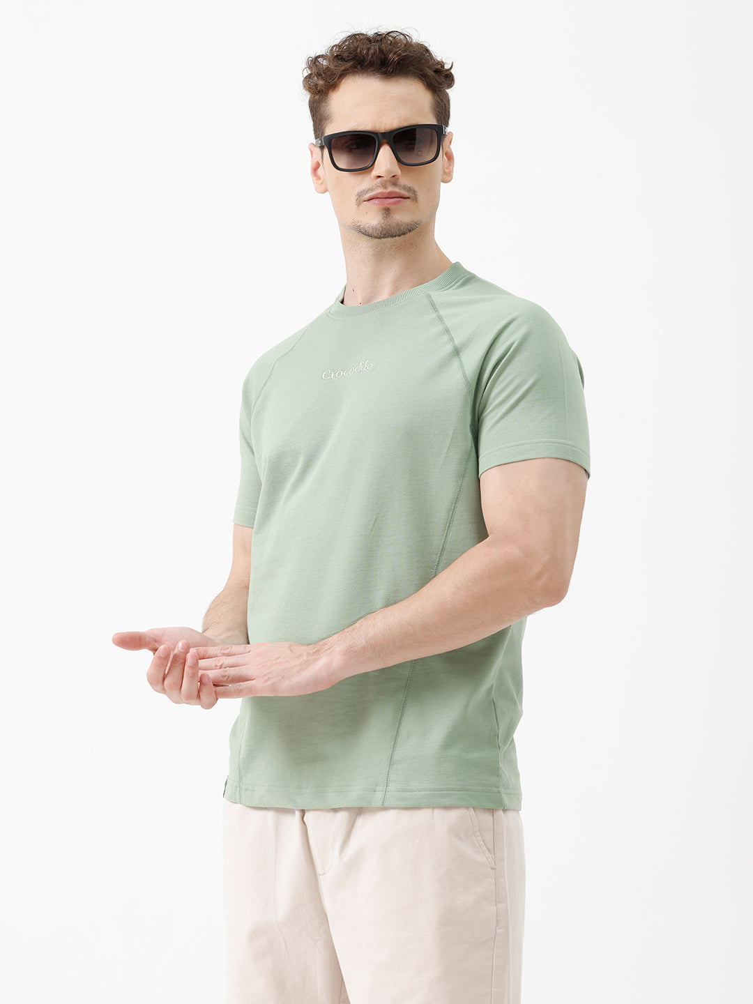 A man wearing a light sage green athletic t-shirt with a small tonal logo on the chest, paired with beige pants, standing against a plain white studio background.