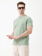 A man wearing a light sage green athletic t-shirt with a small tonal logo on the chest, paired with beige pants, standing against a plain white studio background.
