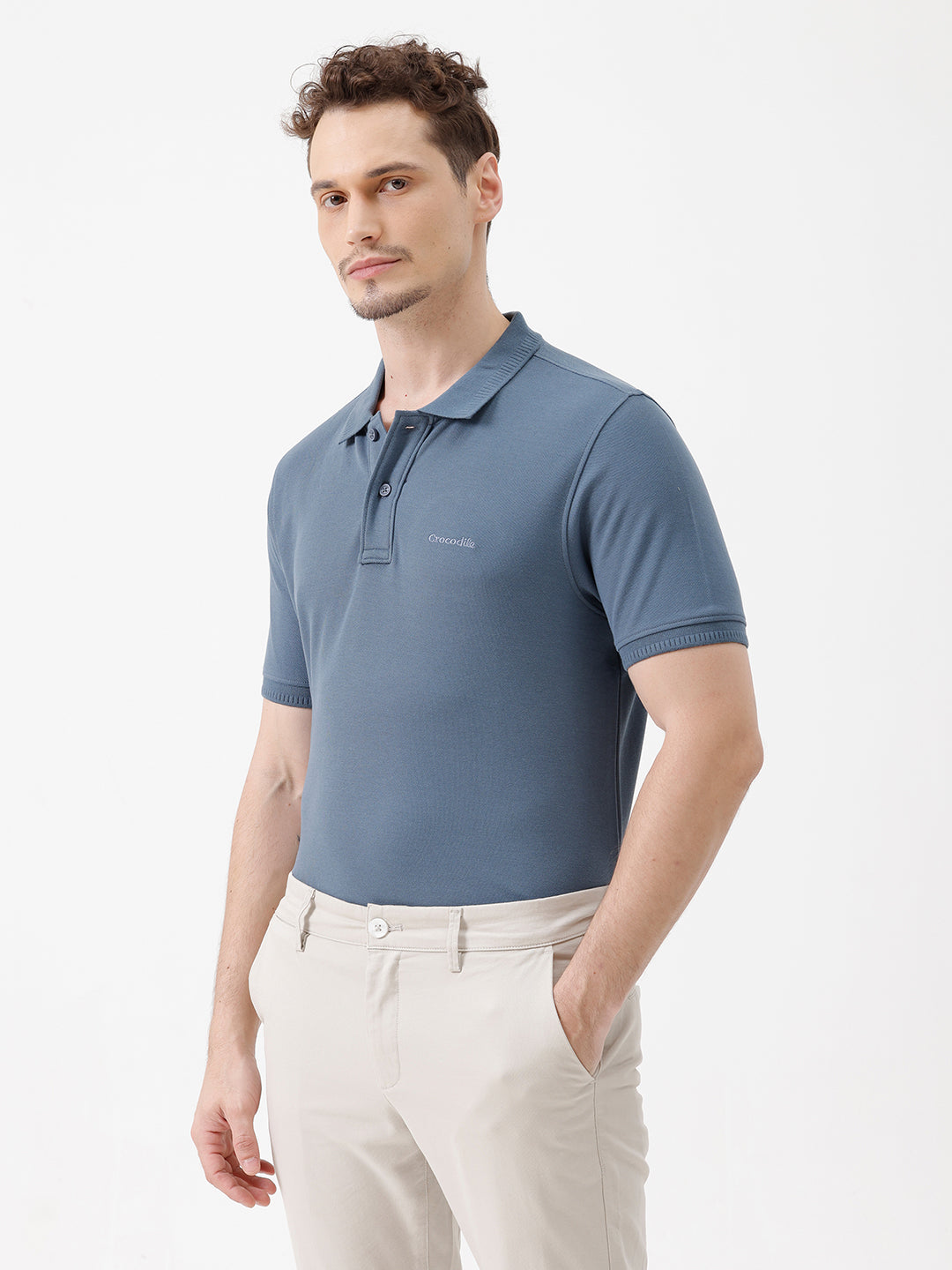 A man wearing a slate blue short-sleeve polo shirt and beige trousers, standing against a plain white studio background.