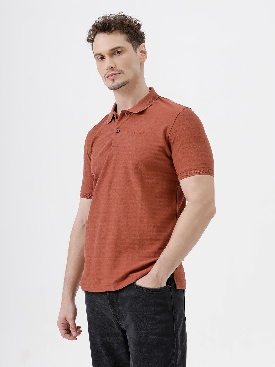 A man wearing a rust-orange short-sleeve polo shirt with a subtle horizontal stripe pattern, paired with dark jeans, standing against a plain white studio background.