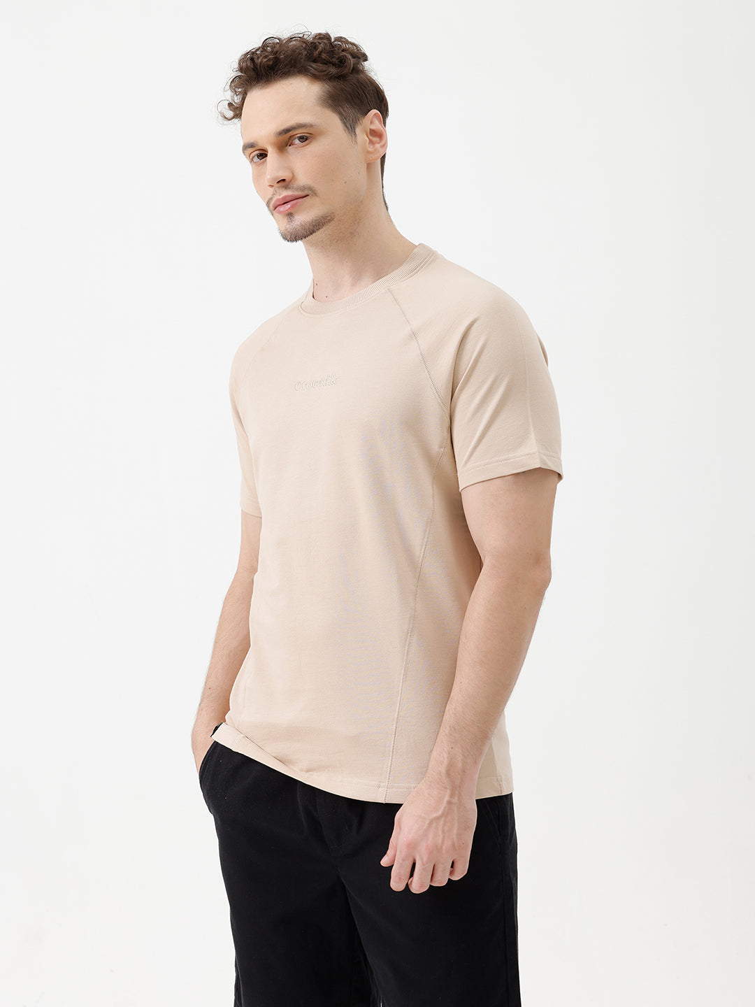 A man wearing a light beige short-sleeve t-shirt with a subtle tonal logo on the chest, paired with black pants, standing against a plain white studio background.