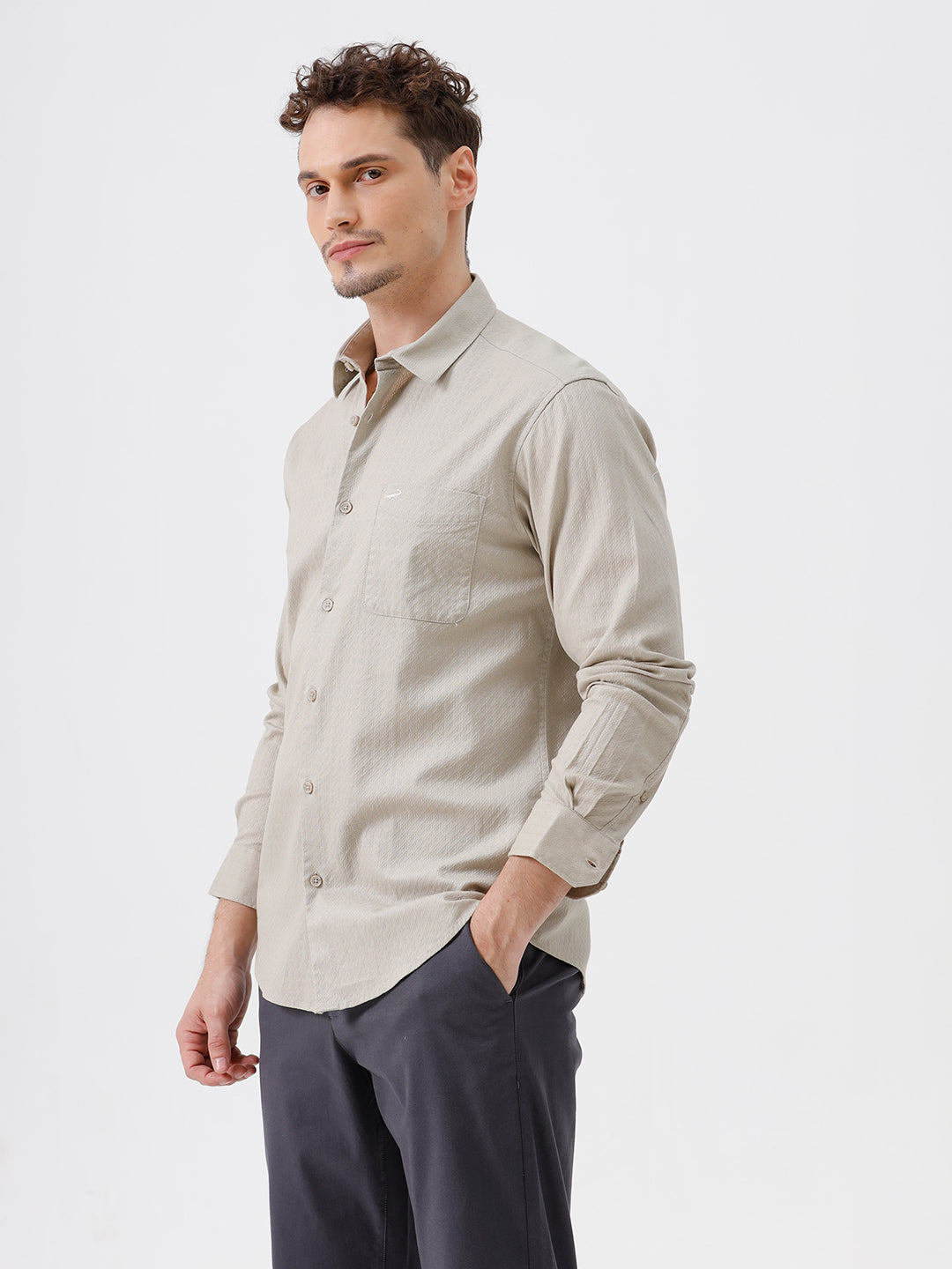 A man wearing a light beige long-sleeve button-down shirt with a chest pocket, paired with dark gray trousers, standing against a plain white studio background.