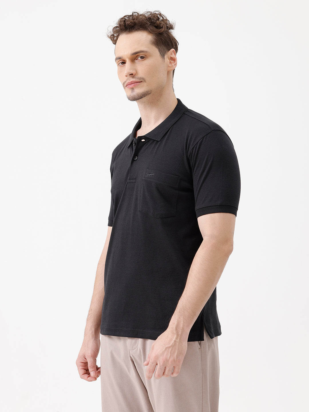A man wearing a black short-sleeve polo shirt with a single chest pocket, paired with beige trousers, standing against a plain white studio background.
