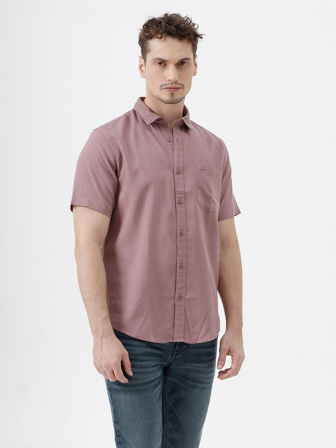 A man wearing a short-sleeve button-up shirt in dusty rose pink with dark blue jeans, standing against a plain white studio background.