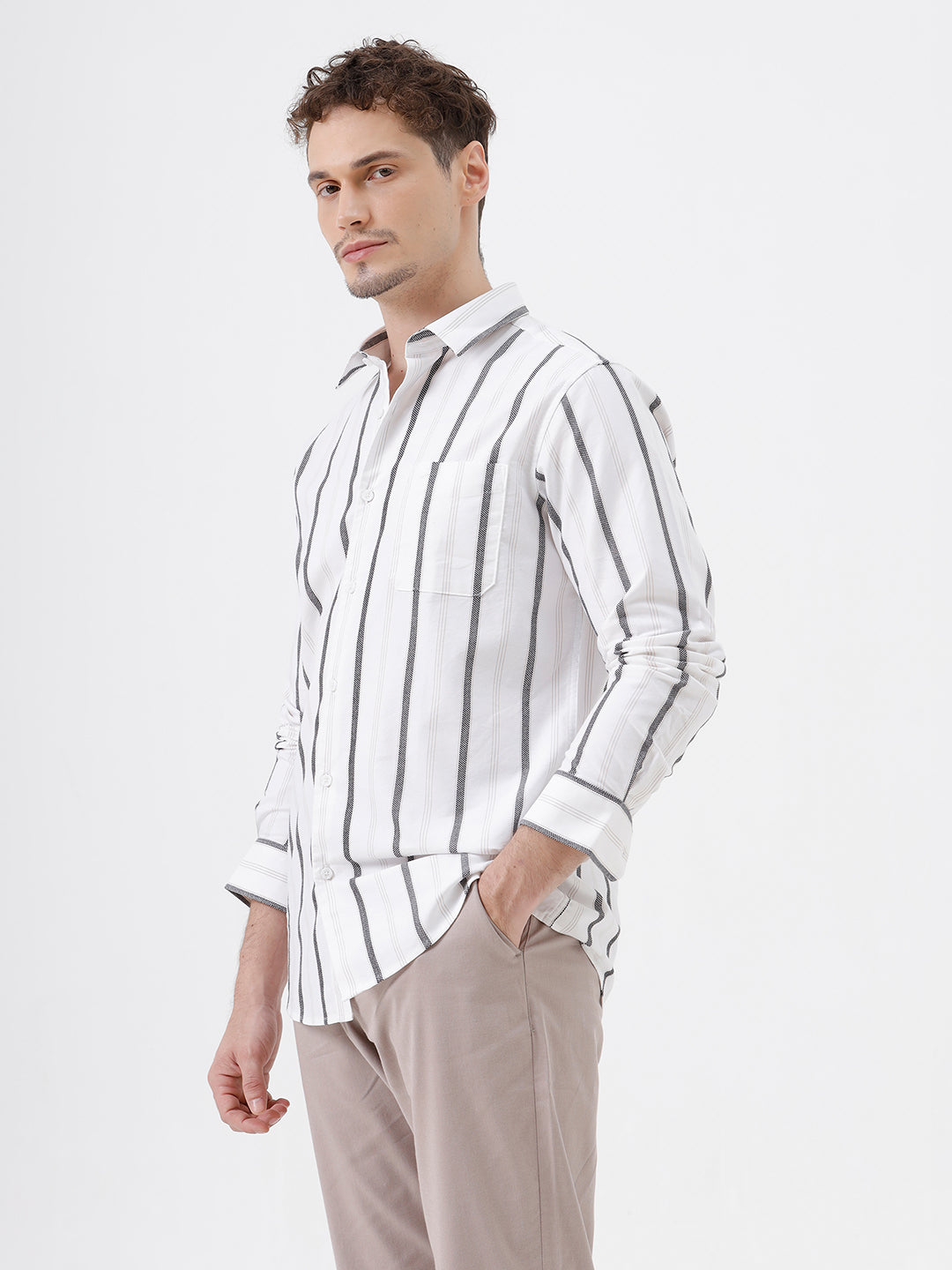 A man wearing a white long-sleeve button-up shirt with vertical gray stripes and beige trousers, standing against a light gray studio background.